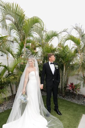 Elegant couple in wedding attire pose in tropical garden setting with tall palm trees and white wall backdrop.