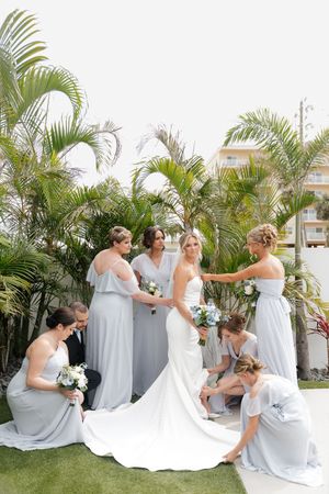 Bridal party in light gray dresses helps arrange bride's long white wedding dress train against tropical palm backdrop.