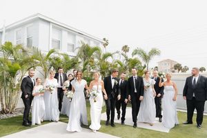 A wedding party in formal attire stands together on a green lawn in front of a white building with palm trees.