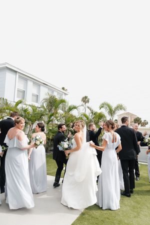 Wedding party in white dresses and black suits gathers on a lush grass lawn beside palm trees at an outdoor venue.