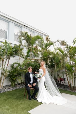 Wedding couple poses in garden setting with tropical palm trees and white building backdrop.