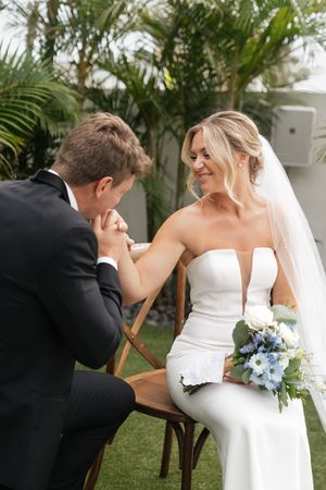 A romantic wedding moment in a garden setting with palm trees as a couple shares an intimate gesture over a bouquet.