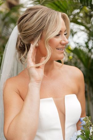 A bride in a white strapless wedding dress and veil smiles during an outdoor ceremony with tropical foliage in background.