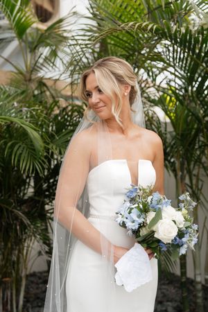 A bride in an elegant white strapless gown holds a blue and white bouquet against a backdrop of tropical palm plants.