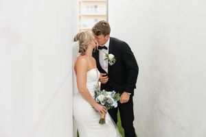 A romantic wedding portrait shows a couple in formal attire with white bouquet against a neutral wall.