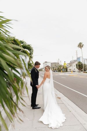 A couple in wedding attire stands on a palm-lined urban sidewalk, with the bride's elegant white gown trailing behind.