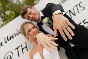 A close-up shot of two hands wearing wedding rings, with palm fronds and text in the background.