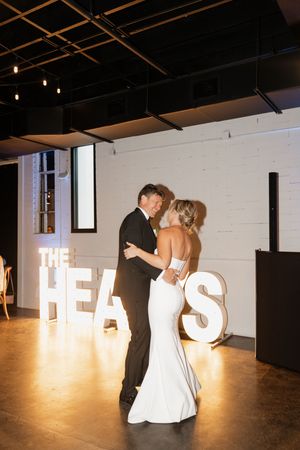 A couple shares their first dance at a wedding reception with illuminated letters spelling HEAPS in the background.