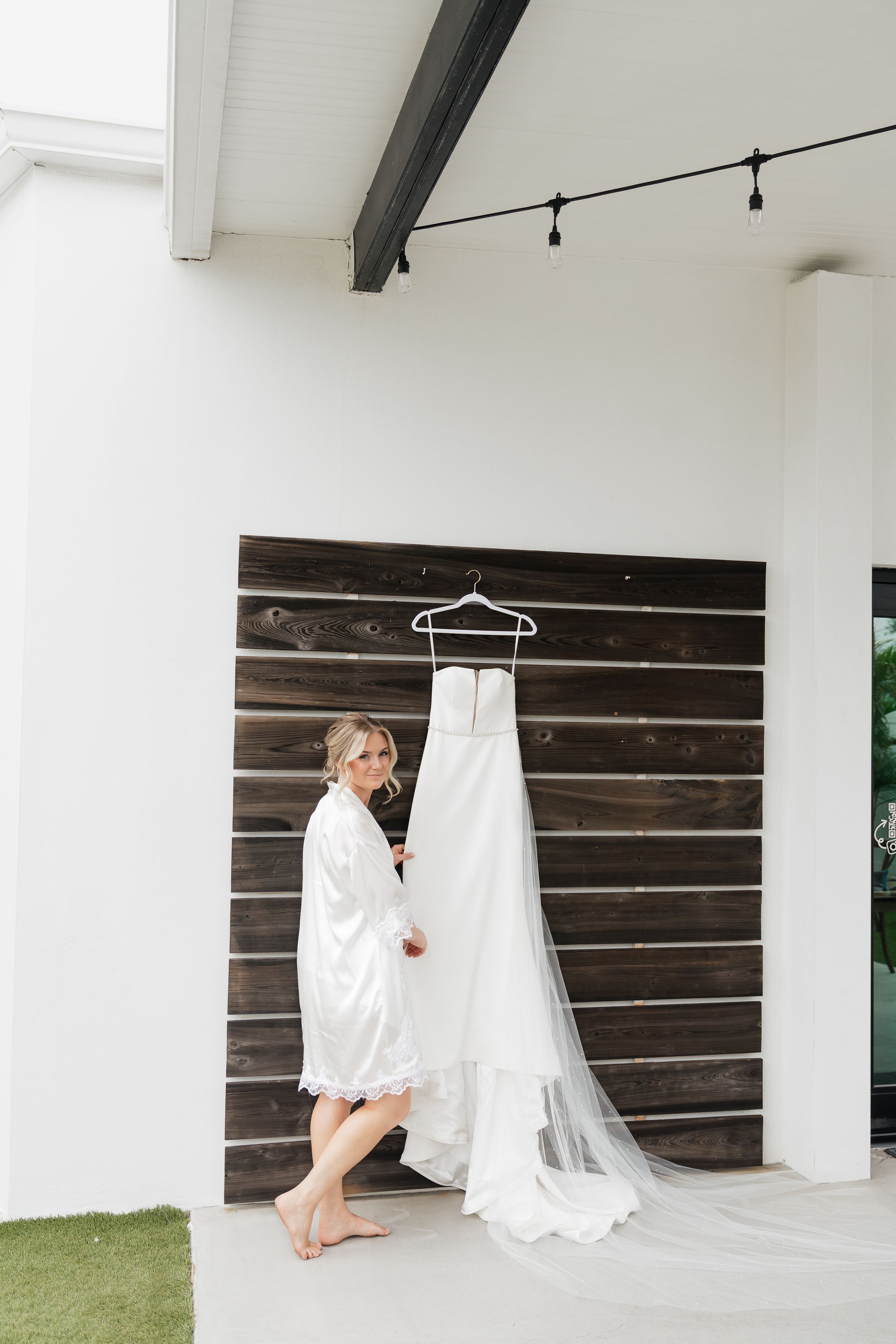 A white wedding dress hangs on a dark wood wall while someone in a robe stands beside it on a covered porch.