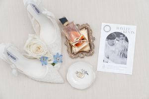 Wedding accessories including white heels, perfume bottle, ring holder, and photo laid out on light beige surface.