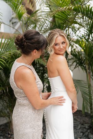 Two women in elegant dresses share a moment together surrounded by tropical palm plants at a formal event.