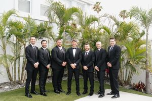 Groomsmen in black tuxedos and bow ties stand in a row against a tropical palm garden backdrop.