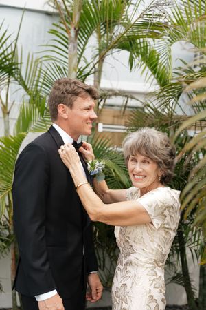 A woman in a cream lace dress adjusts the bow tie of someone wearing a black tuxedo among tropical palm fronds.