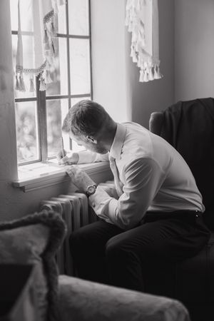 A person sits by a window in a contemplative pose, captured in black and white with soft natural lighting.