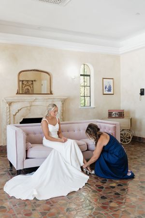 Two people in formal attire sit on a pink couch in an elegant room with terracotta tile floors and arched windows.