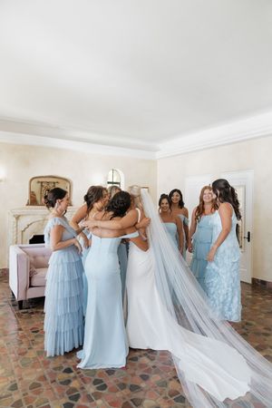 Bridesmaids in light blue dresses gather around a bride's flowing white wedding gown in an indoor setting.
