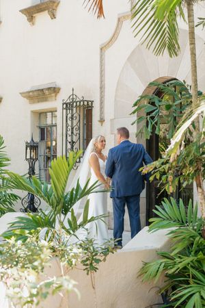 A couple shares a romantic moment among tropical palm plants outside a Mediterranean-style building.