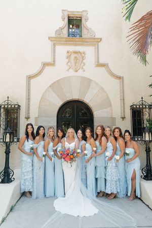 A wedding party in light blue bridesmaid dresses poses against a white Spanish colonial style building exterior.