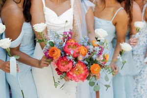 Wedding party in light blue dresses holds colorful bouquets of coral and orange flowers against white backdrop.