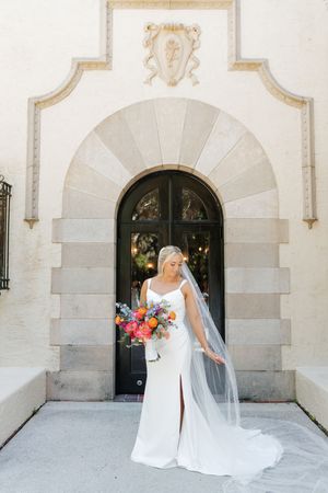 A bride in a flowing white gown poses against an elegant stone archway with ornate architectural details.