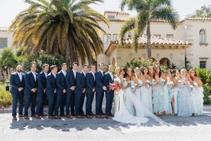 Wedding party in navy suits and light blue dresses lined up in front of a Mediterranean style building.
