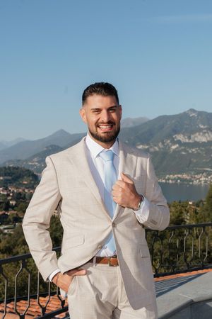 Smiling groom in beige suit posing on a terrace with mountain and lake views.