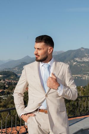 Groom in beige suit and blue tie poses on balcony with mountain lake views.