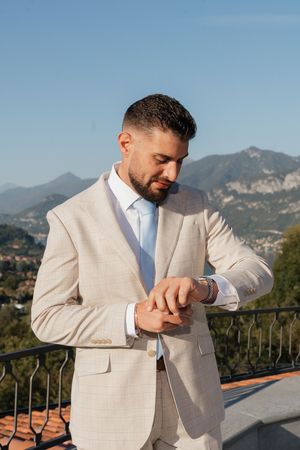 Groom in beige suit adjusting cufflinks on balcony with mountain backdrop on wedding day.