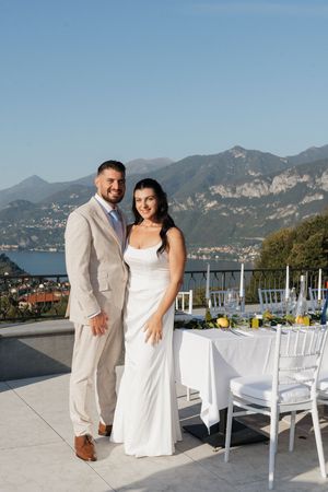 Smiling bride and groom posing together beside decorated reception tables with mountain lake views.