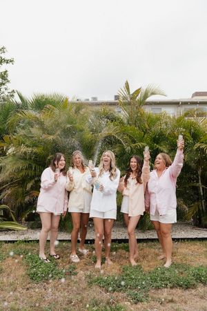 A group of friends in light-colored outfits pose joyfully together outdoors against a backdrop of tropical palm trees.