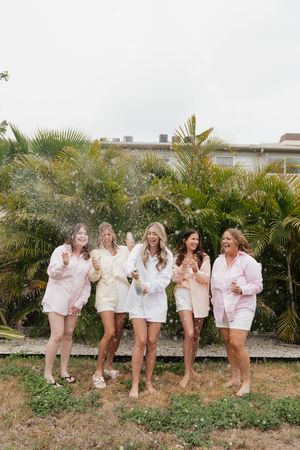 Five women in matching pink and white outfits laugh together while standing in front of tropical palm trees.