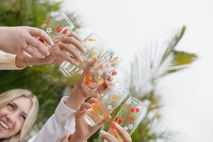 Group of friends cheering with colorful tropical drinks outdoors under palm trees in the sunlight.