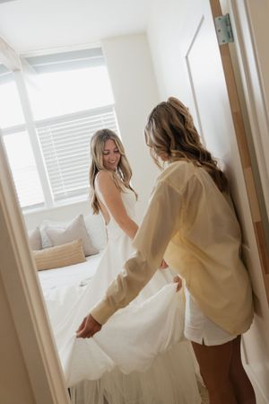 Two women in a bright white bedroom preparing a bed with fresh linens and bedding in natural sunlight.