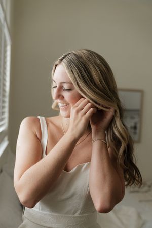 Natural light portrait of woman in white tank top smiling with eyes closed by window in soft morning light.