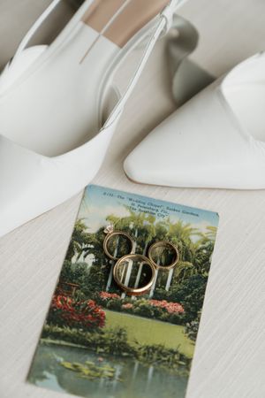 Wedding rings rest on a vintage postcard beside elegant white bridal shoes on a light surface.
