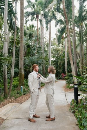 Two people in cream-colored suits adjust each other's attire while standing on a pathway surrounded by tall palm trees.