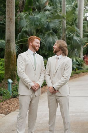 Two people in matching light beige suits share a joyful moment together on a palm-lined walkway.