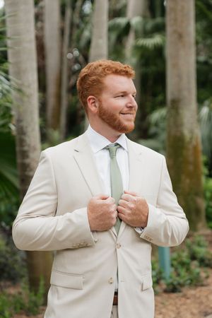 A groom in a cream-colored suit and sage green tie stands in a tropical garden setting with palm trees.