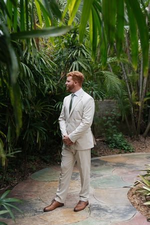 A person in a cream-colored suit stands thoughtfully on a stone path surrounded by lush tropical garden foliage.