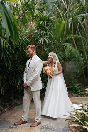 A couple in tropical garden wearing light beige and white wedding attire holding colorful bouquet.