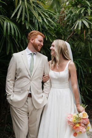Newlywed couple in elegant cream-colored attire share a joyful moment against lush tropical palm fronds.