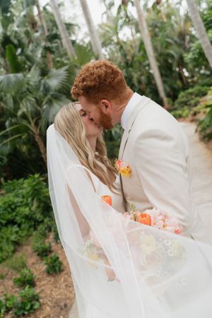A romantic wedding moment captured in a lush tropical garden setting with peach flowers and flowing white veil.