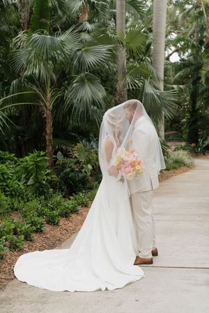 A couple shares a romantic moment under a white veil surrounded by tropical palm trees and lush greenery.