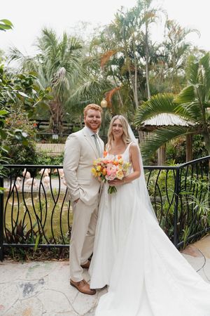 A wedding couple stands together in a lush tropical garden setting with palm trees and ornate metal railings in the background.