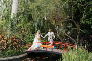 Two people pose on a red Japanese garden bridge surrounded by lush tropical foliage and bamboo trees.