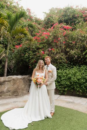 A couple poses for a wedding photo in front of blooming red bougainvillea and palm trees on a garden patio.