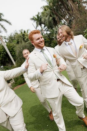 A joyful wedding party wearing cream colored suits dances and celebrates on a green lawn surrounded by palm trees.