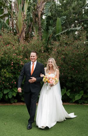 A wedding ceremony takes place in a lush tropical garden setting with banana trees and flowering bushes in the background.