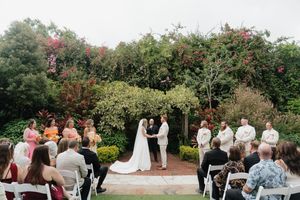 Outdoor garden wedding ceremony with guests seated on white chairs and blooming flowers in the background.