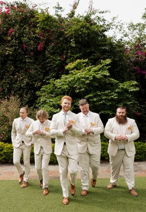 A group of men in cream colored suits walk together on grass with flowering bushes in the background.